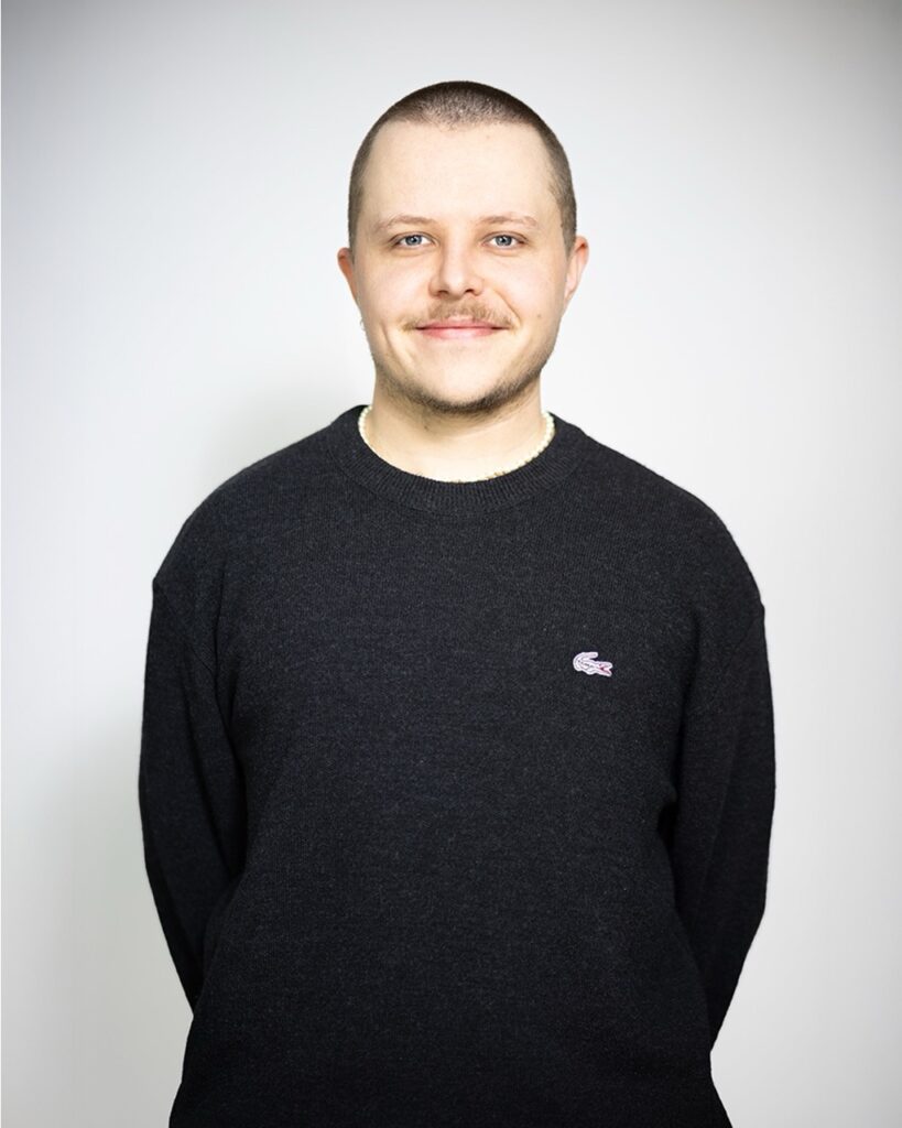 Mann mit rasiertem Kopf und Schnurrbart, trägt einen dunkelgrauen Pullover mit einem kleinen Krokodil-Logo, lächelt leicht in die Kamera.