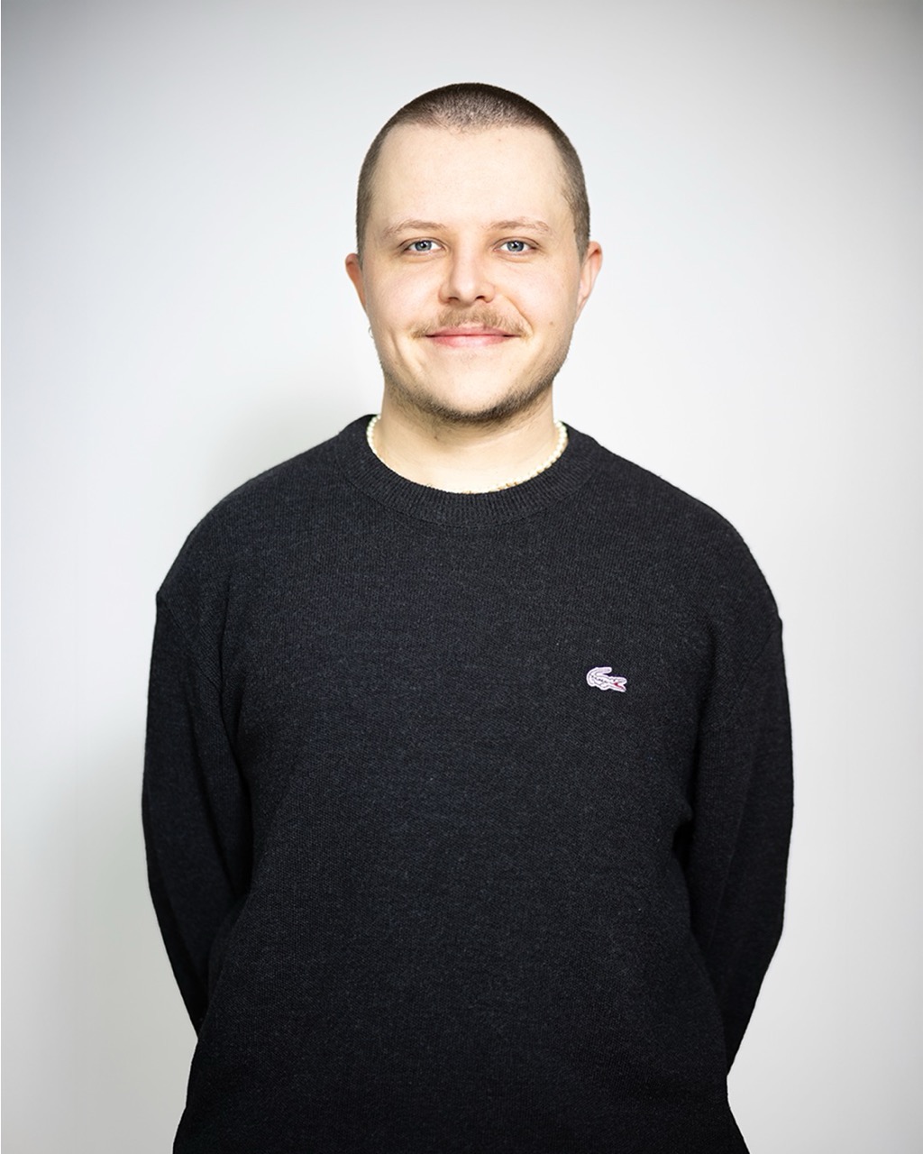 Mann mit rasiertem Kopf und Schnurrbart, trägt einen dunkelgrauen Pullover mit einem kleinen Krokodil-Logo, lächelt leicht in die Kamera.