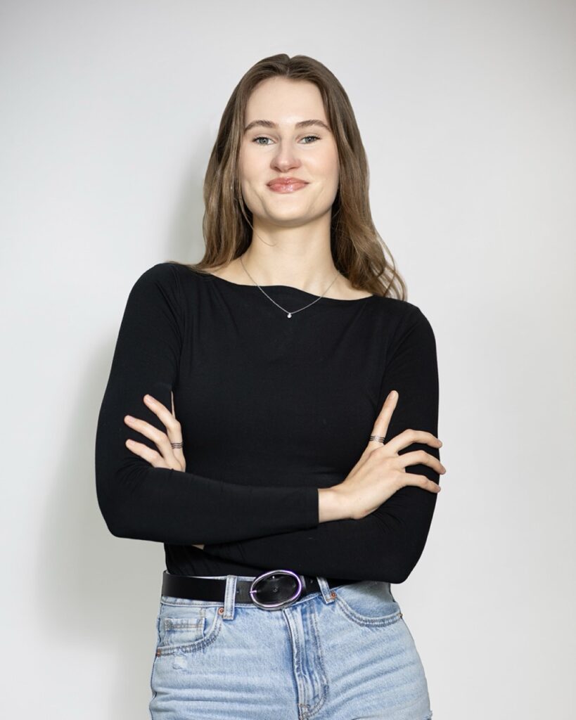 Junge Frau mit langen, braunen Haaren, trägt ein schwarzes Langarmshirt und helle Jeans, steht mit verschränkten Armen und einem selbstbewussten Lächeln vor hellem Hintergrund.