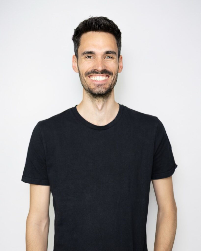 Mann mit kurzen, dunklen Haaren, Dreitagebart und strahlendem Lächeln, trägt ein schwarzes T-Shirt und steht offen und freundlich vor hellem Hintergrund.