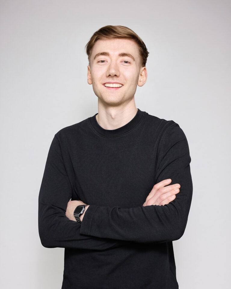 Junger Mann mit kurzem, braunem Haar und hellem Teint, trägt ein schwarzes Langarmshirt und eine digitale Armbanduhr, steht mit verschränkten Armen und lächelt freundlich.