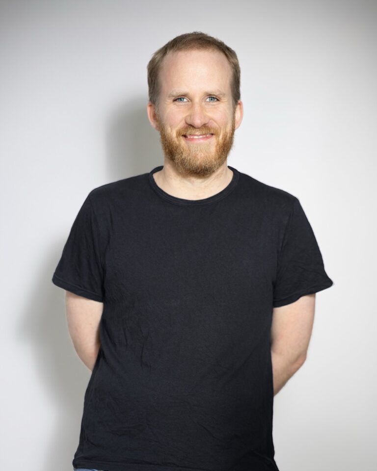 Mann mit kurzem, rötlich-blondem Haar und Vollbart, trägt ein schwarzes T-Shirt und steht mit entspanntem Lächeln vor hellem Hintergrund, die Arme hinter dem Rücken verschränkt.