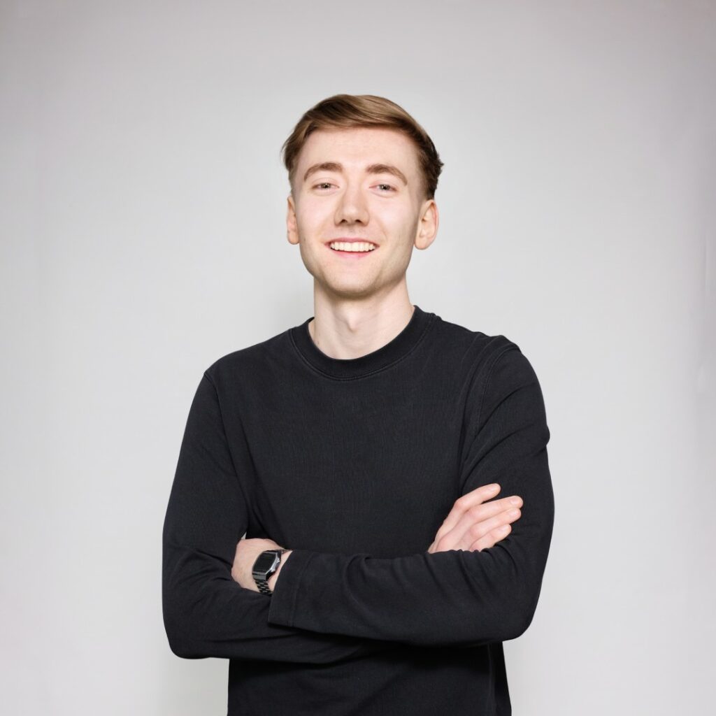 Oskar Eder, Werkstudent SEO - Junger Mann mit kurzem, braunem Haar und hellem Teint, trägt ein schwarzes Langarmshirt und eine digitale Armbanduhr, steht mit verschränkten Armen und lächelt freundlich.