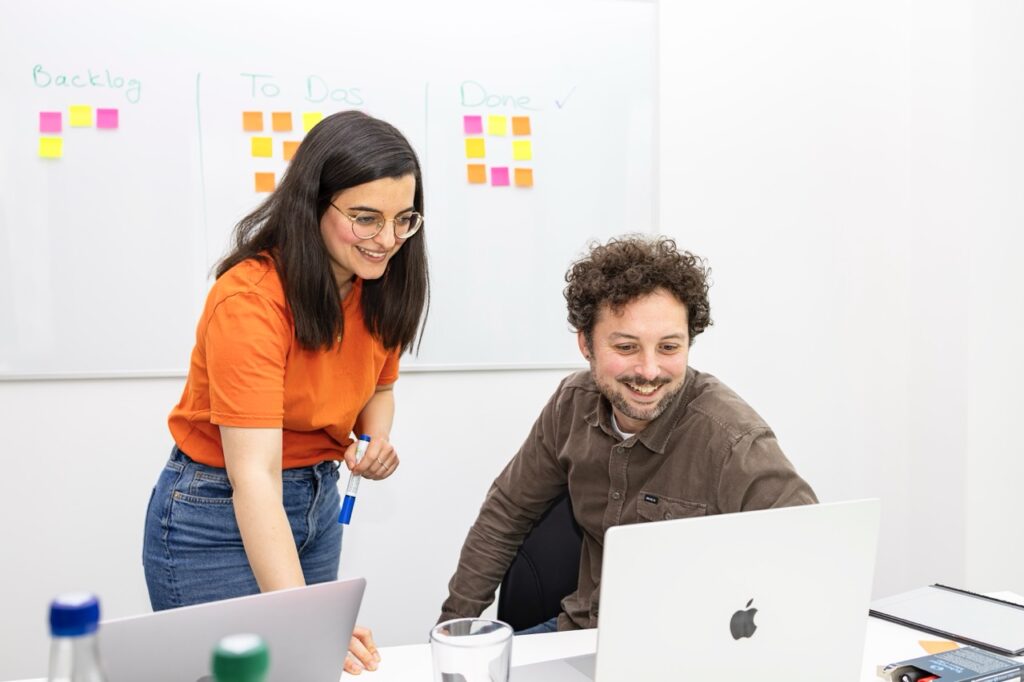 Zwei Kolleg:innen arbeiten gemeinsam vor einem Laptop in einem modernen Büro. Die Frau im orangefarbenen Shirt steht lächelnd neben dem sitzenden Mann, der ein braunes Hemd trägt – beide schauen auf den Bildschirm. Im Hintergrund ist ein Whiteboard mit den Spalten „Backlog“, „To Dos“ und „Done“ sowie bunten Haftnotizen zu sehen, was auf ein agiles Projektmanagement hindeutet.