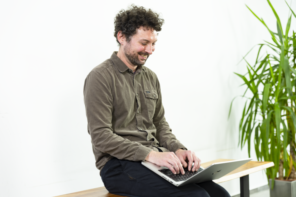 Ein Mann mit lockigem Haar und Bart sitzt auf einer Holzbank und arbeitet lächelnd an einem Laptop. Er trägt ein braunes Hemd und eine dunkle Hose. Der Hintergrund ist weiß und minimalistisch, die Szene wirkt freundlich und fokussiert.