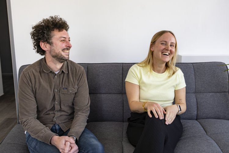 Zwei fröhliche Personen, ein Mann und eine Frau, sitzen nebeneinander auf einem grauen Sofa. Beide lachen herzlich. Der Mann trägt ein braunes Hemd und Jeans, die Frau ein hellgelbes T-Shirt und schwarze Hose. Die Szene wirkt freundlich und entspannt vor einem weißen Hintergrund.