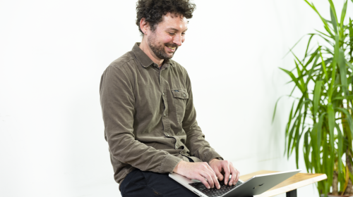 Ein Mann mit lockigem Haar und Bart sitzt auf einer Holzbank und arbeitet lächelnd an einem Laptop. Er trägt ein braunes Hemd und eine dunkle Hose. Der Hintergrund ist weiß und minimalistisch, die Szene wirkt freundlich und fokussiert.