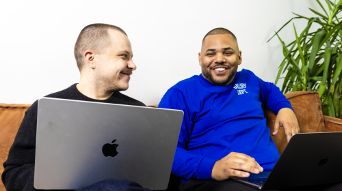 Zwei Männer sitzen nebeneinander auf einem braunen Sofa, lachen und arbeiten jeweils an ihren Laptops, während sie sich ansehen.